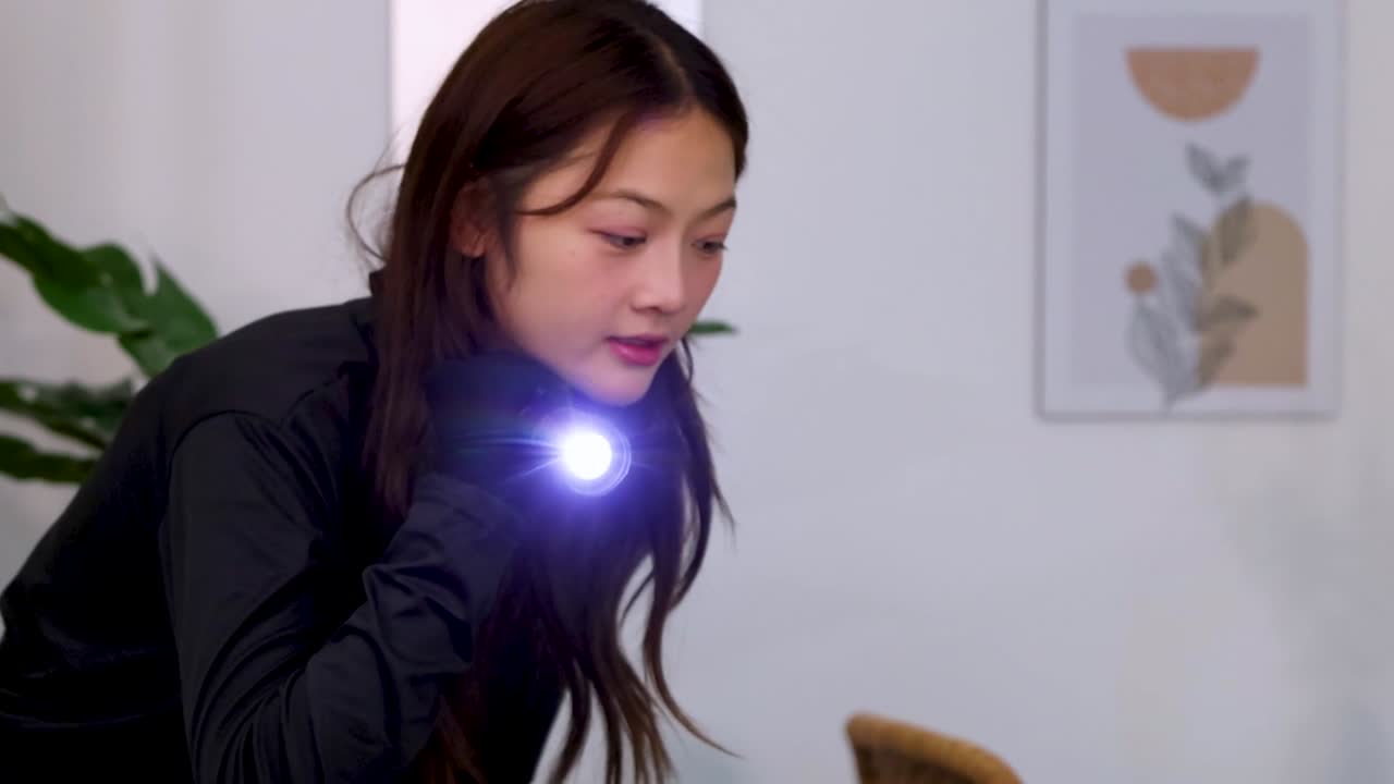 A woman in black clothing cautiously searches a modern room using a handheld flashlight beam
