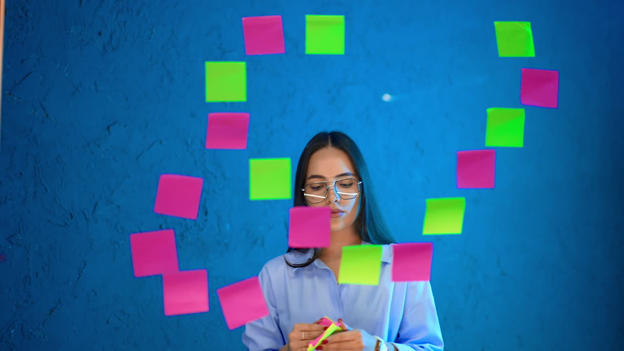 Woman notes in office. A woman in glasses uses sticky notes to organize ideas on a blue wall in a modern office setting during the day