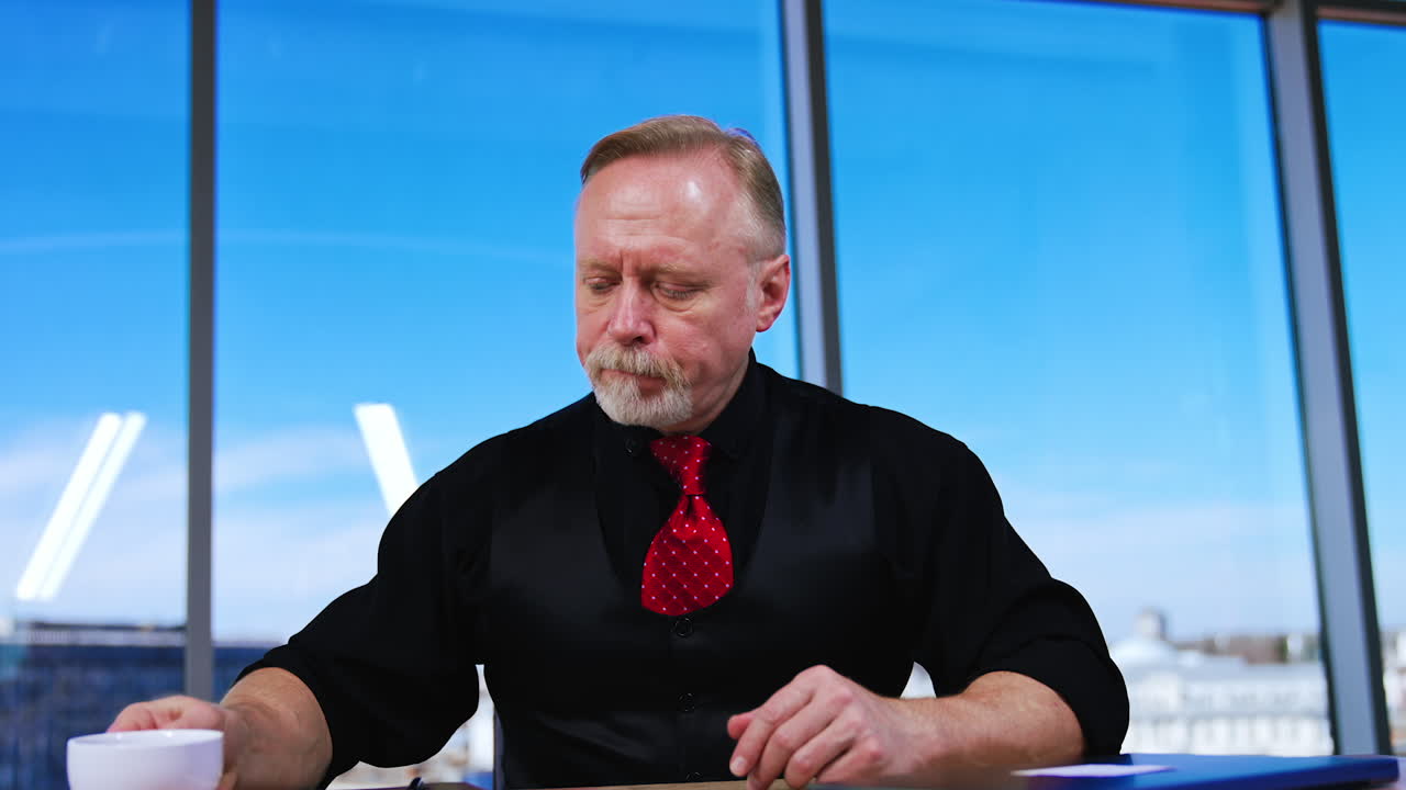 Old man in black clothes and red tie drinks coffee in office. Businessman opens his laptop and starts work.