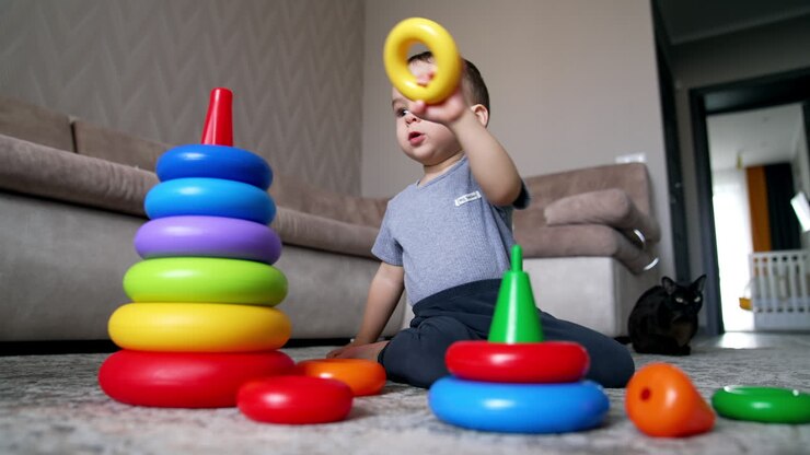 Calm serious baby boy plays on the floor. Kid constructs toy pyramids stacking the rings. Black cat sits beside watching the child. Low angle view.