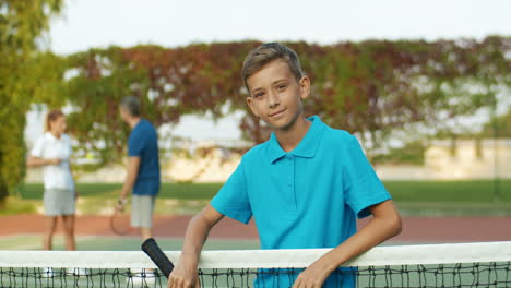 portrait of a cute teen boy with tennis racket in hand leaning on net and smiling cheerfully at the camera 1