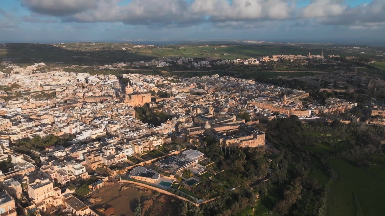 Vista aerea di una città storica con edifici bianchi e beige, campanili di chiese e campi verdi circostanti sotto un cielo parzialmente nuvoloso.