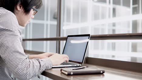 young asian business man using laptop computer in working space with smartphone and notebook on wooden desk. male hand typing on laptop keyboard. freelance lifestyle in digital age concept.