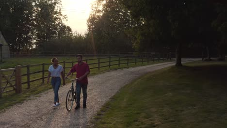 Drohnenaufnahme-Eines-Romantischen-Paares,-Das-Bei-Sonnenuntergang-über-Die-Landstraße-Läuft-Und-Das-Fahrrad-Schiebt