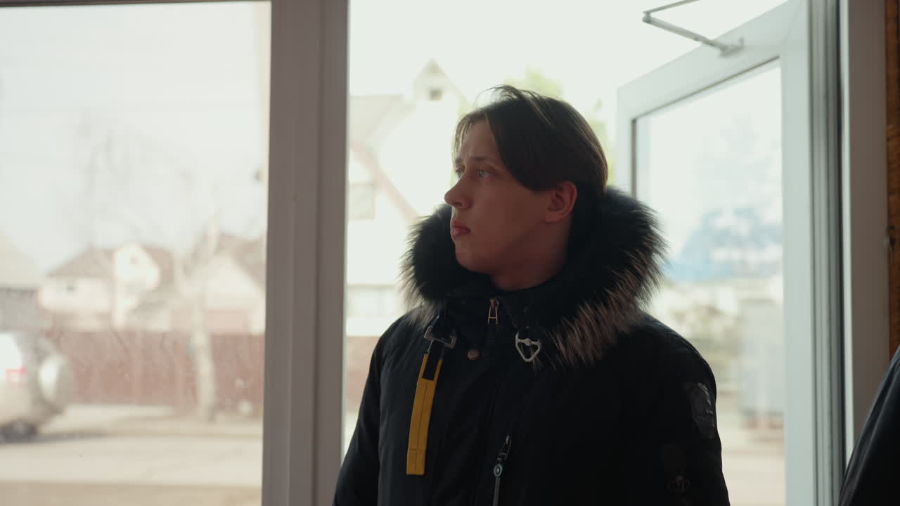 Two men step inside through glass door, one wearing sunglasses and backpack, other in winter coat with fur hood, both looking around interior space with suburban houses and street visible behind them