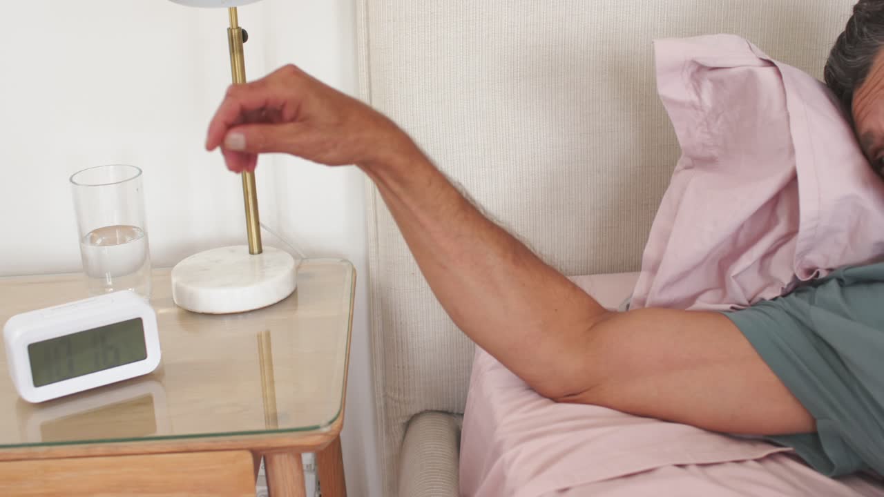 Mature couple in bed with alarm ringing, man waking and reaching across pink sheets silencing clock