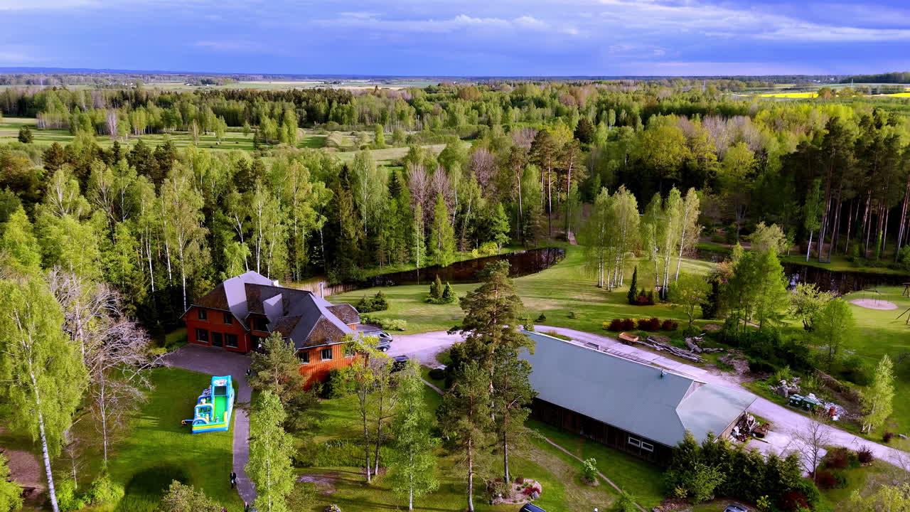 Spacious rural estate featuring houses, trees, driveways, and open green space, nestled in dense forest with distant farmland and moody spring skies above