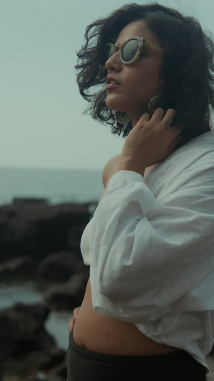 Vertical shot of a South Asian woman in sunglasses and white shirt by the rocky seashore