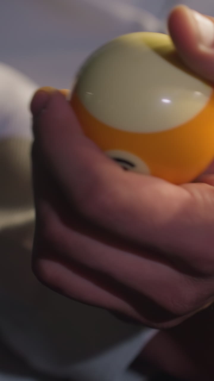 Primer plano de una mano en una camisa blanca, sin que se vea la cara, sujetando un libro abierto mientras hace rodar una bola de billar en una mano. Gesto concentrado, iluminación melancólica e intensa concentración.