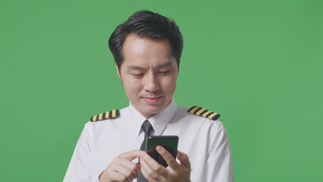 녹색 스크린 배경 스튜디오에 서 있는 동안 미소 짓고 스마트폰을 사용하는 아시아 남성 조종사의 클로즈업