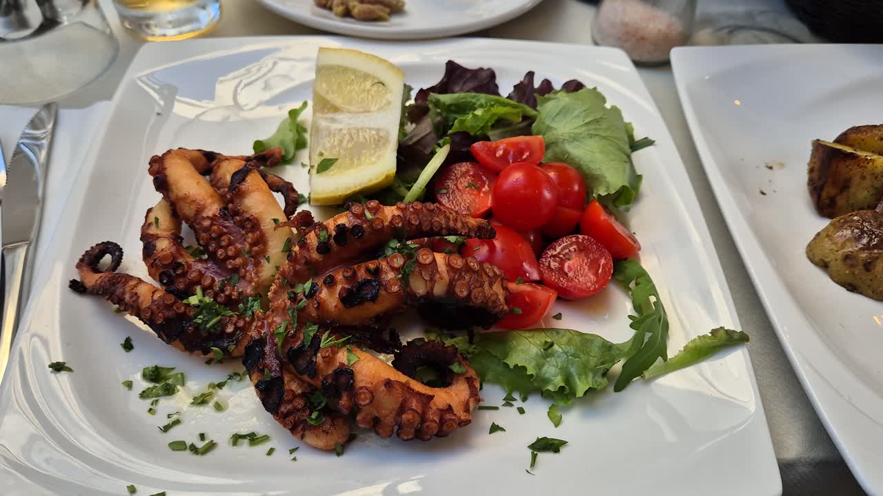 pulpo a la parrilla en un plato blanco con ensalada verde, tomates y limón