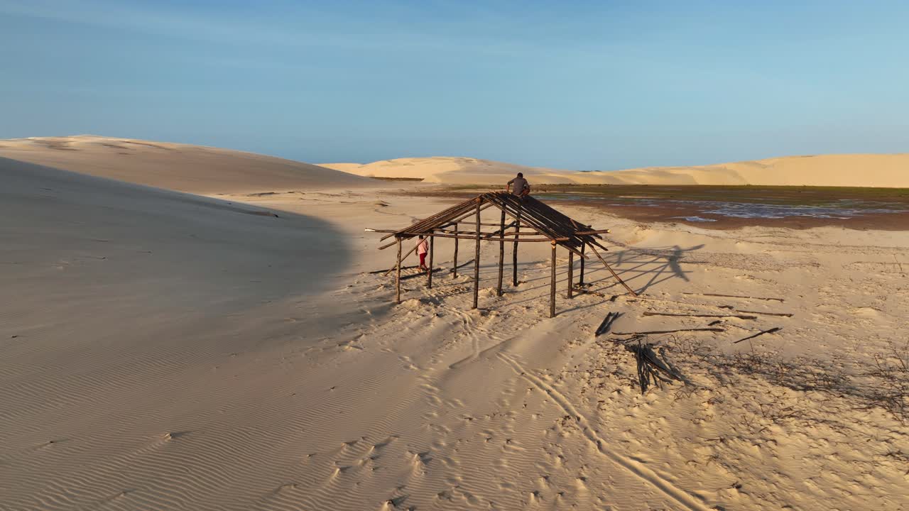 Men work on wooden shelter frame amid sand dunes near Parnaíba River in Araioses, Maranhão, Brazil. Aerial orbiting