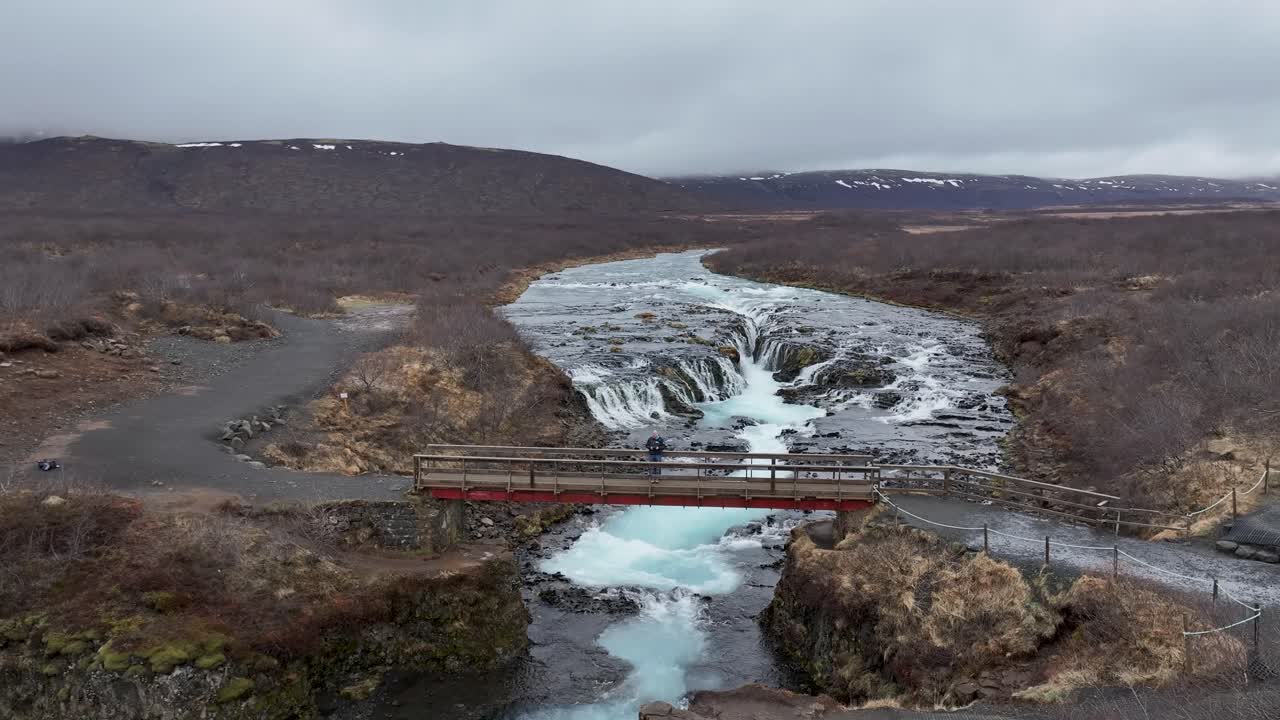 Amidst Iceland's rugged splendor, a man stands at the edge of a bridge, drone in hand, unlocking the secrets of the land and revealing a symphony of natural wonders, Waterfall Bruararfoss