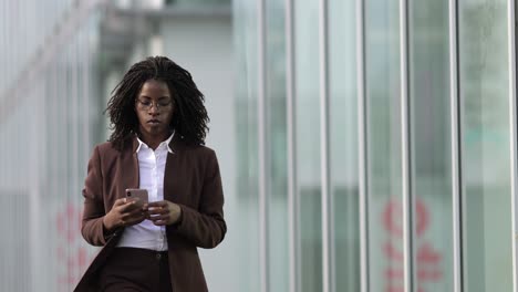 Ernsthafte-Afroamerikanische-Frau,-Die-Beim-Spaziergang-Ihr-Smartphone-Benutzt