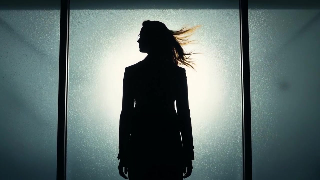 Donna in piedi con blazer che sente una brezza fuori campo che le solleva i capelli, proprio davanti a un alone di vetro smerigliato.
