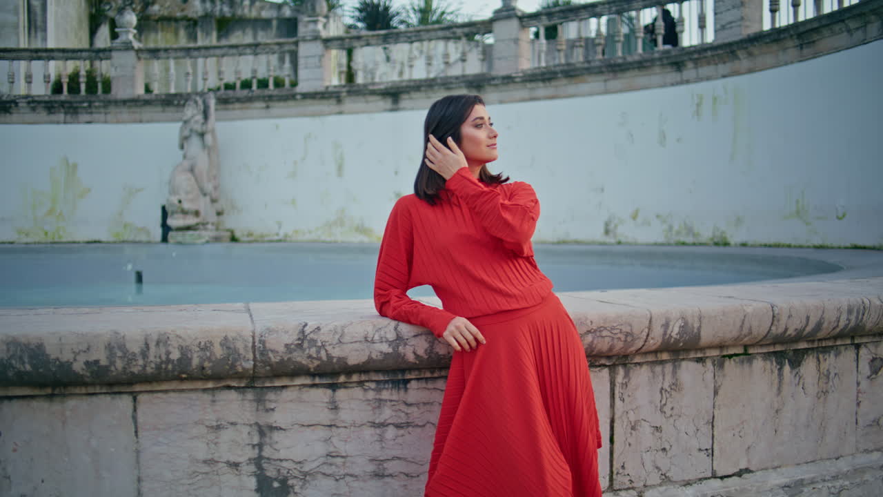 Chic lady looking camera at water fountain. Woman in red dress adjusting hair