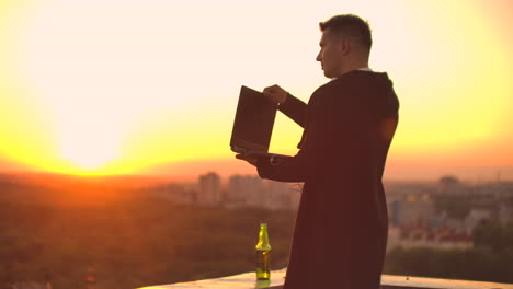 un corredor de bolsa independiente está de pie en un techo al atardecer con una computadora portátil y escribe en un teclado con los dedos mirando el paisaje de la ciudad desde la vista de un pájaro.