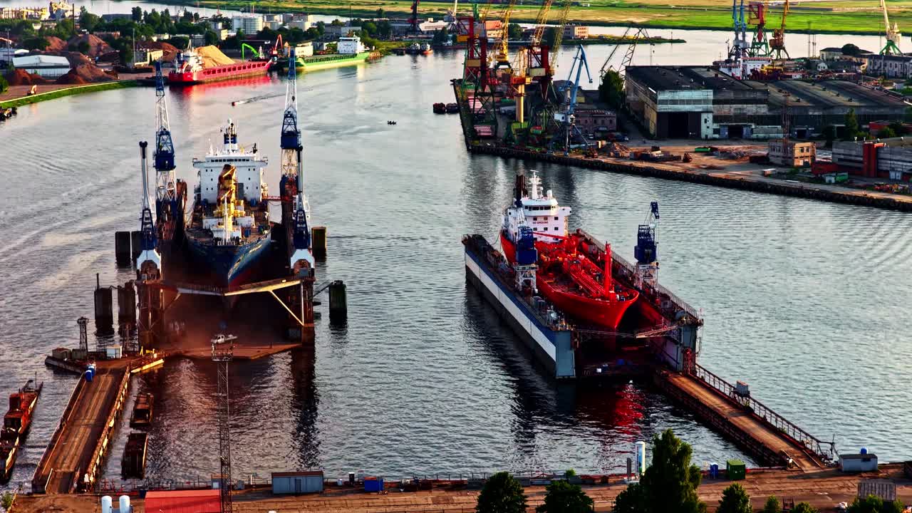 Två stora fartyg ligger i flytande torrdockor längs en livlig industriell vattenväg, omgivna av kranar, reparationsplattformar och hamnbyggnader, marinteknik och fartygsunderhåll