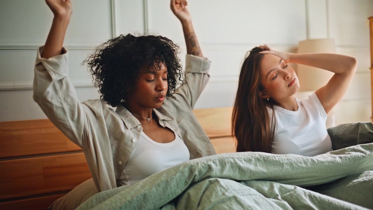 Lesbian couple waking up stretching morning bed. Multiracial women home bedroom.