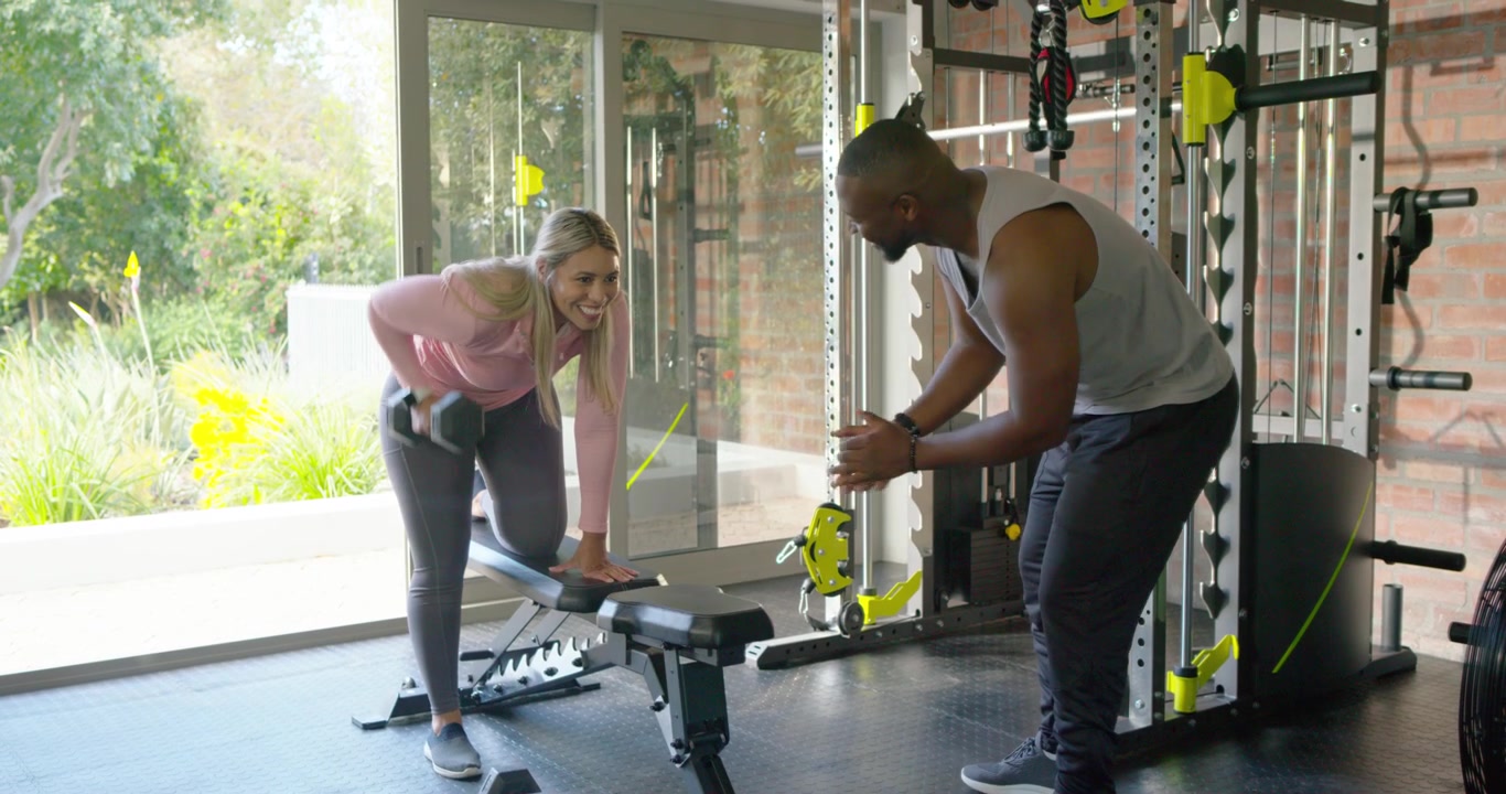 African American trainee on bench picking dumbbell doing one-arm rows while trainer coaching in gym