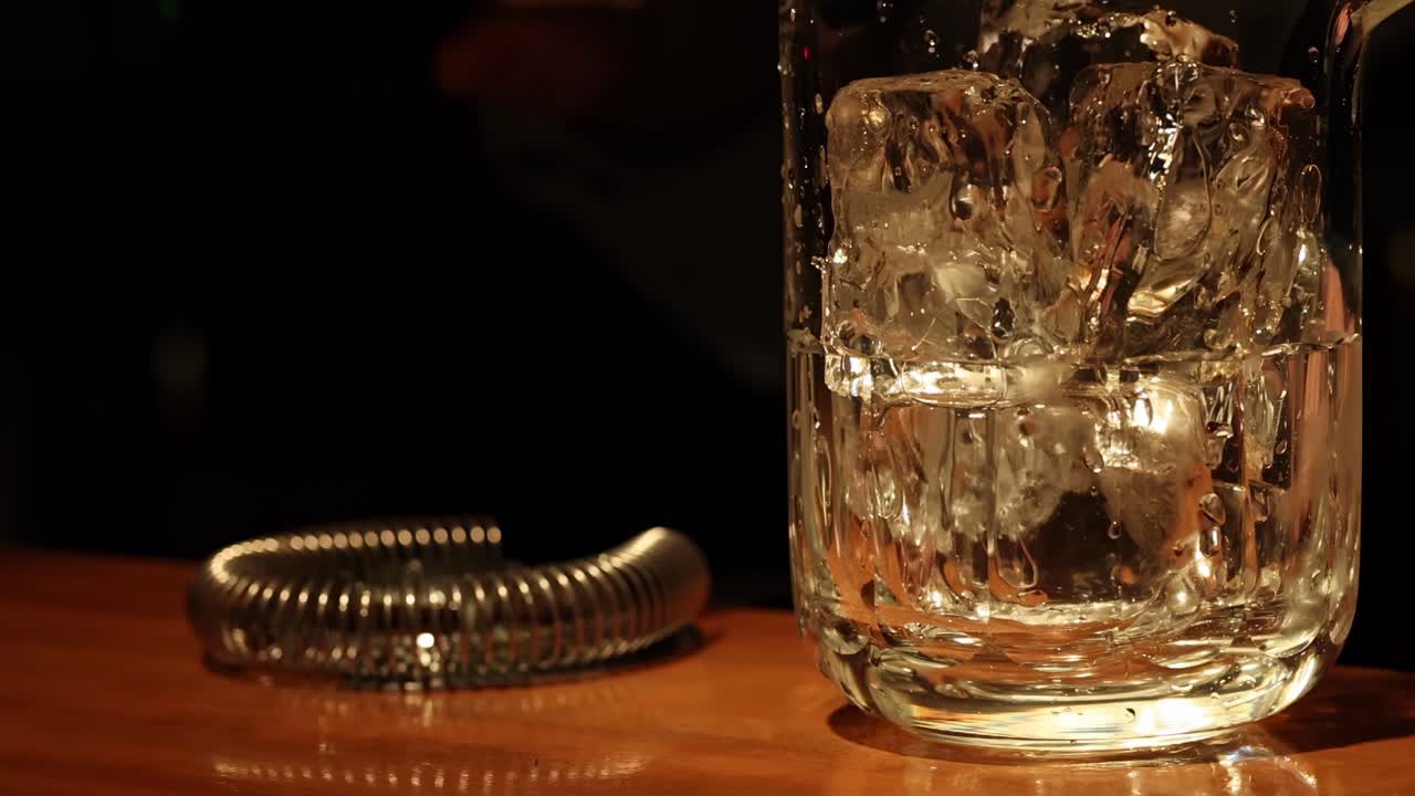 A close-up of whiskey being stirred with ice in a glass beside a metal strainer.