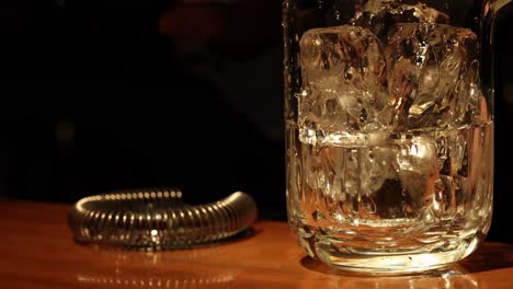 a close-up of whiskey being stirred with ice in a glass beside a metal strainer.