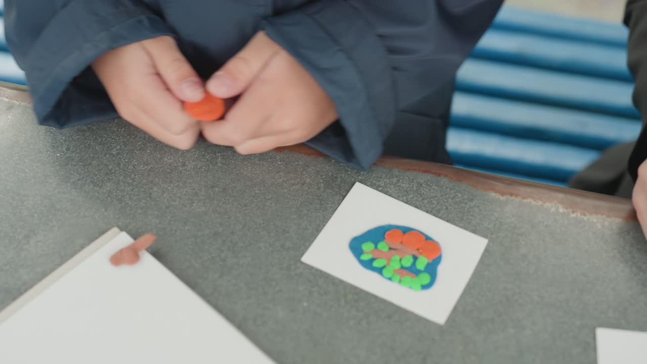 Child hands crafting colorful clay flower on bench, student molding orange petal with fingers, small paper card with bright floral design, jacketed child at school table preparing craft to take back