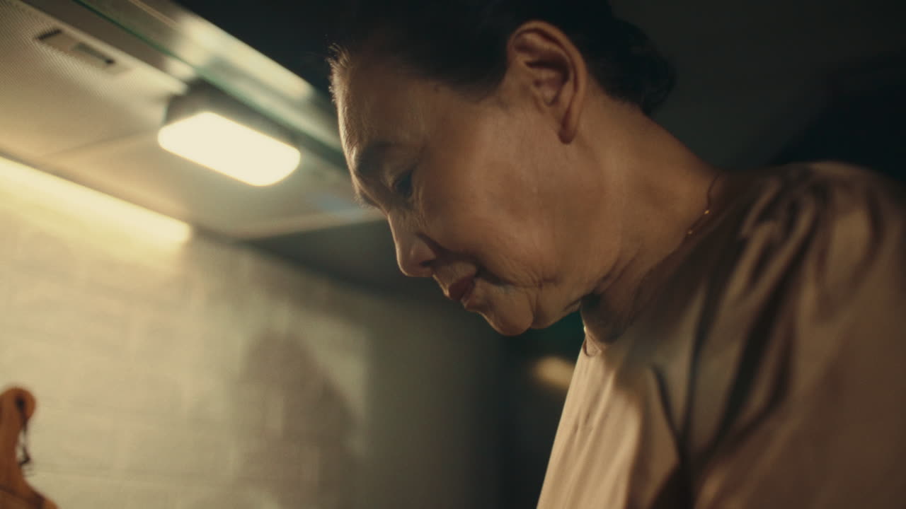 Sad older woman lost in thought, standing in her kitchen, dimly lit environment