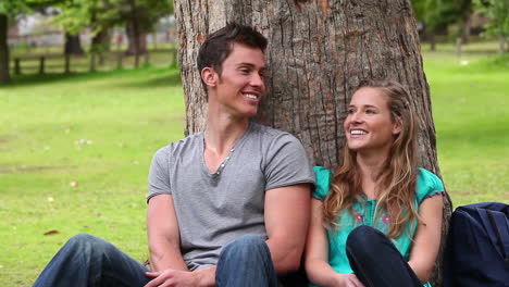 Two-friends-smiling-as-they-look-at-each-other-while-sitting-against-a-tree