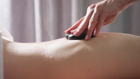 close view of massage therapist gently gliding two smooth black stones on client back during professional therapy session, captured in soft lighting with gray curtain background for calm ambiance