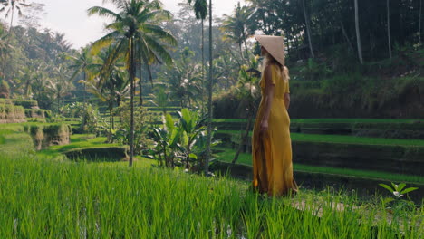 woman walking in rice paddy wearing yellow dress with conical hat exploring lush green rice terrace in cultural landscape exotic vacation through bali indonesia discover asia