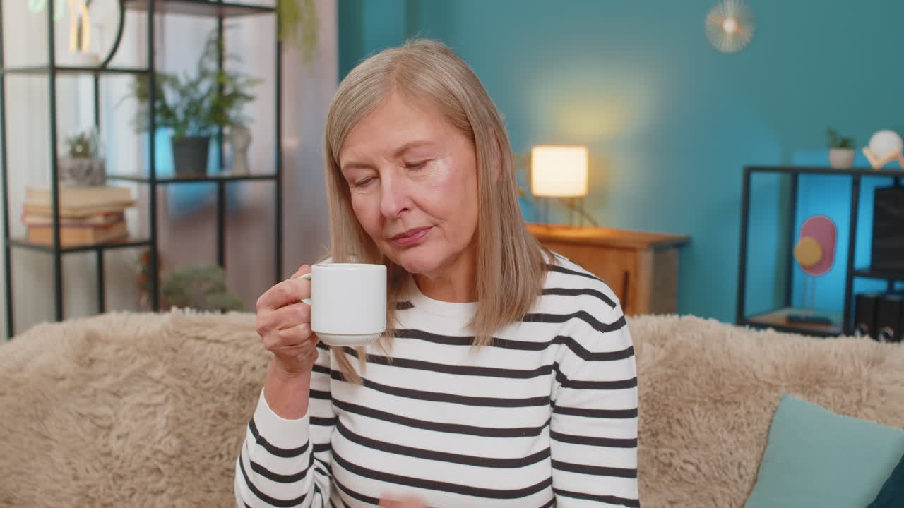 Happy mature woman raising coffee cup smiling in greeting or gratitude cheers gesture on home sofa