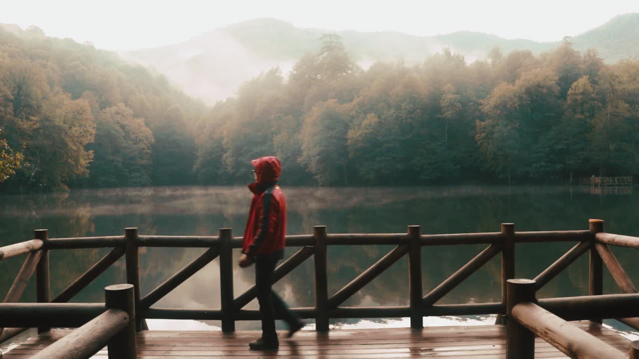 Anonymous male tourist in red raincoat walking on wooden platform at calm Lake Como against trees and mountain. The peaceful scene highlights natural beauty and serene surroundings.