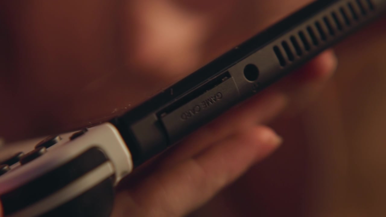 Tight handheld close view of electric shaver guided along facial skin, fingers holding matte plastic handle while metal blade housing makes light contact with visible stubble, with warm indoor light