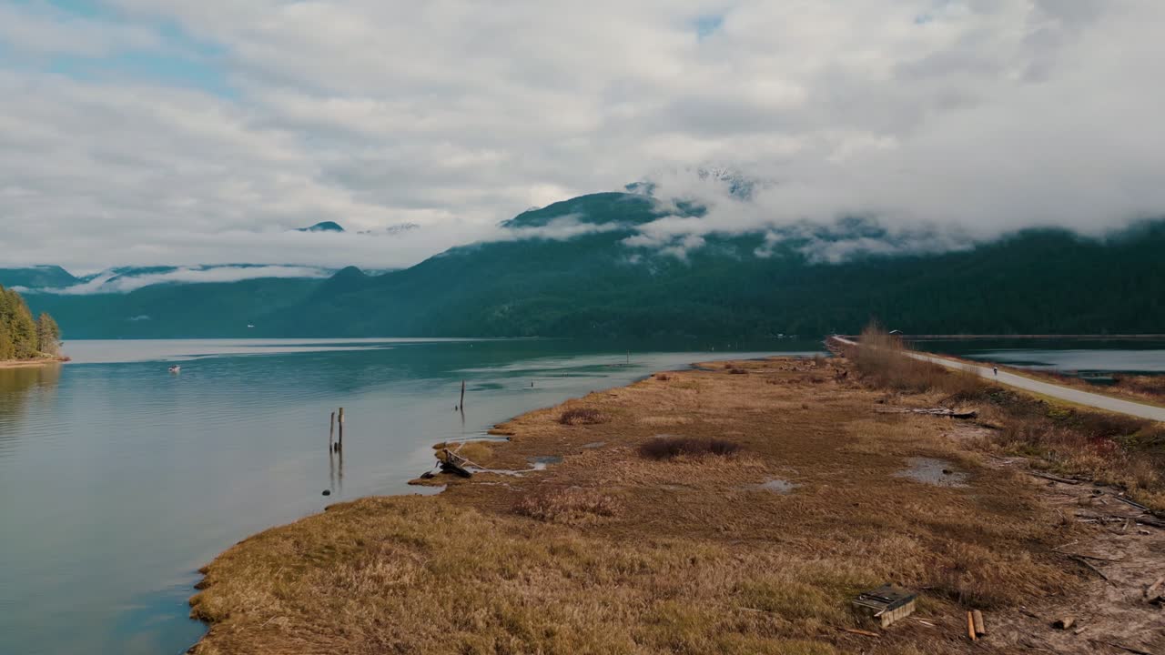 un camino de grava en la naturaleza que conduce a un tranquilo camino pacífico del río rodeado de agua hierba marrón amarilla, niebla niebla montañas de color verde oscuro nubes en el otoño aéreo que se acerca a maple ridge pitt meadows bc
