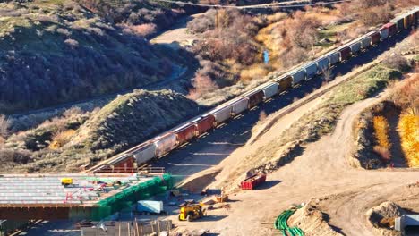 Aerial---Train-Passing-Below-a-Bridge-Construction-in-Bluffdale-Utah,-Tracking-Telephoto-Lens