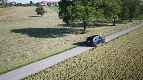 A-Volvo-car-drives-on-a-small-road-near-a-village-in-the-swiss-countryside,-Vaud,-drone-shot