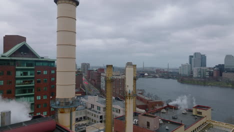 Fly-around-chimneys-of-factory-on-riverbank.-Revealing-aerial-view-of-urban-boroughs-along-river.-Boston,-USA