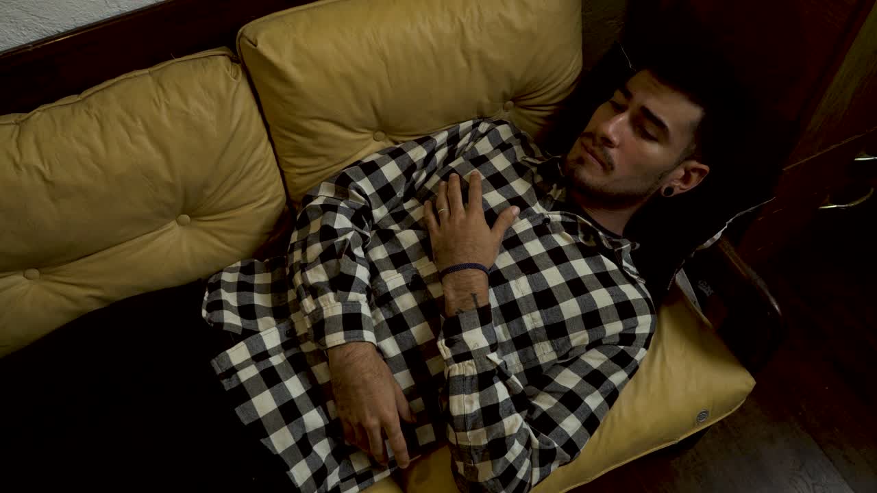 High-angle view of a tired young man in a plaid shirt lying down on a couch, relaxing at home, and falling into a deep sleep