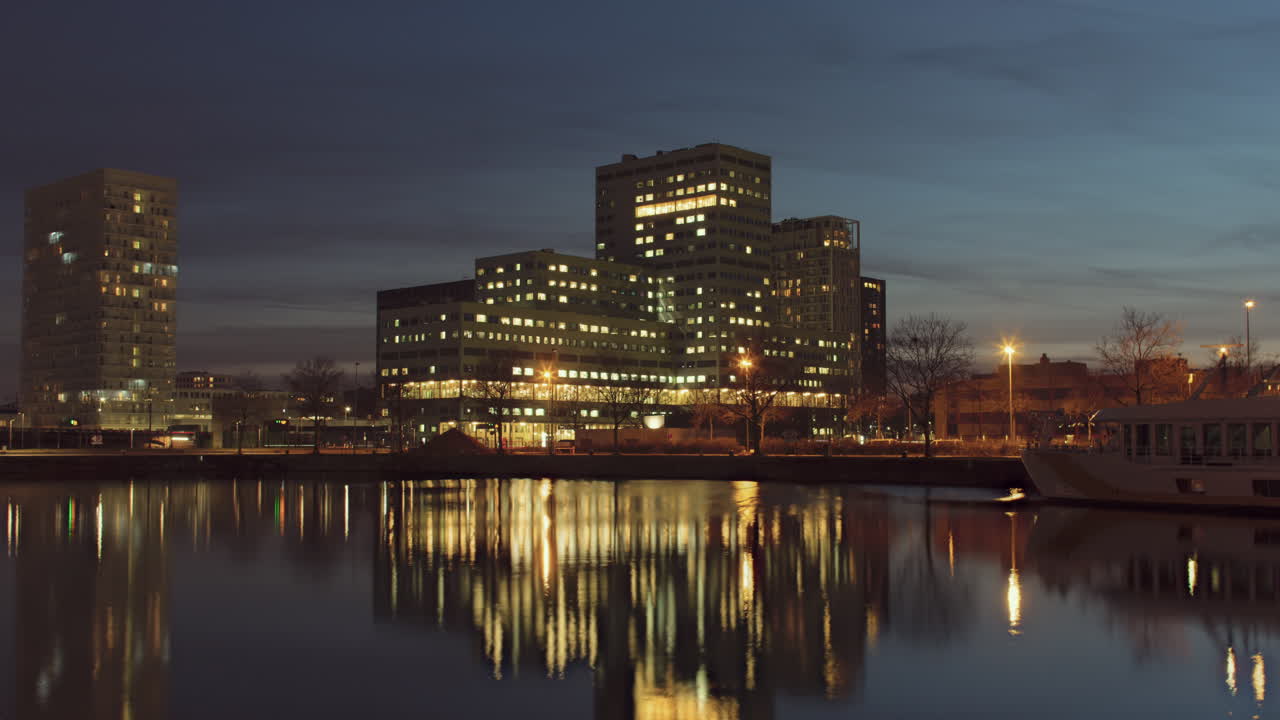 Timelapse at dusk of a modern cityscape with illuminated buildings reflecting in the calm waters, featuring the ZNA Cadix hospital in Antwerp, Belgium