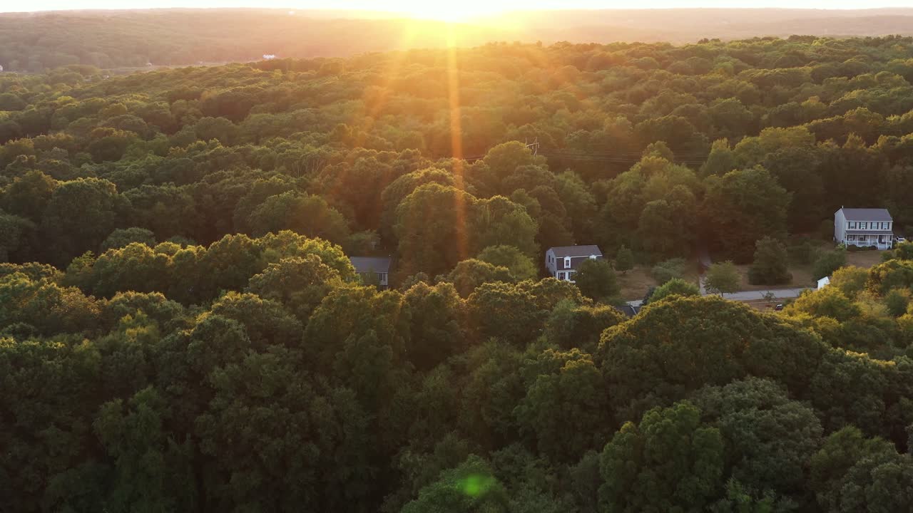 fast aerial flight heads toward the bright summer sunset over rural homes in a forested area of the Northeastern USA, with large lens flare