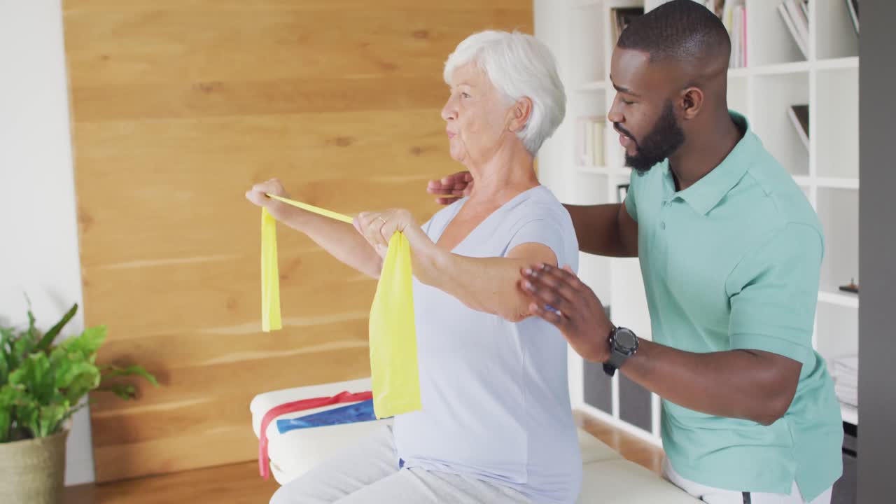 Video of happy caucasian senior woman exercising with african american male physiotherapist