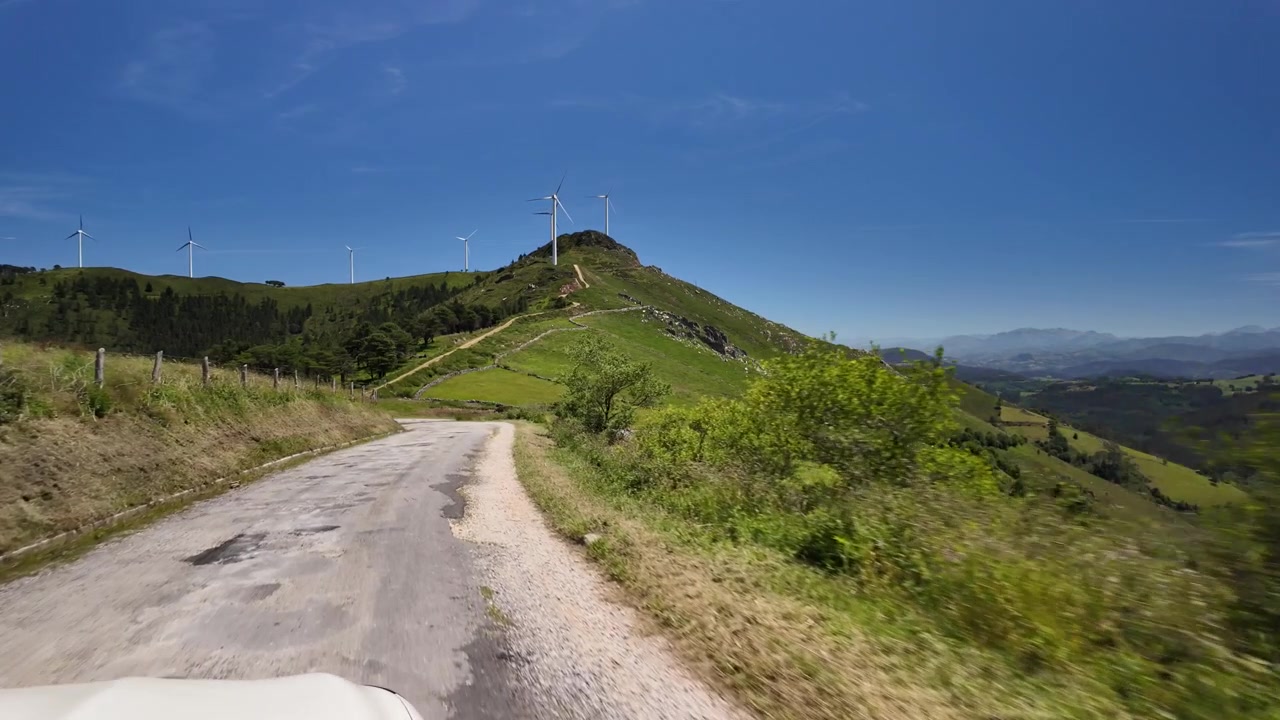 Vista dal conducente su una strada di montagna che conduce verso pale eoliche distanti.