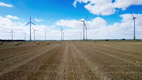 Arriba,-Una-Hilera-De-Turbinas-Eólicas-Gira-Con-Gracia-En-El-Campo-De-Un-Granjero-De-Lincolnshire,-Recién-Cosechado,-Con-Fardos-De-Heno-Dorados-En-El-Frente