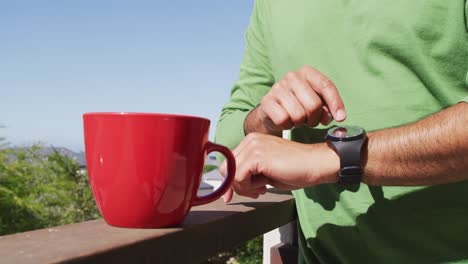 Mixed-race-man-having-a-cup-of-coffee-checking-smartwatch-on-balcony-in-sun