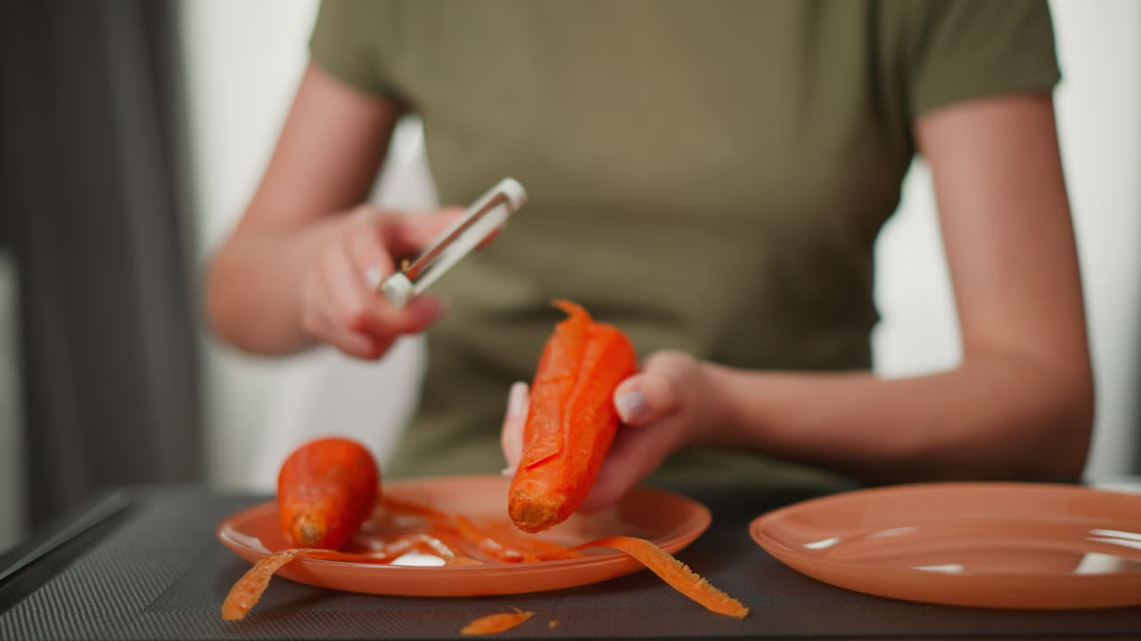 Portrait view of woman in green top holding carrot over orange plate while peeling with handheld peeler, carrot skin scattered on surface, calm indoor environment with soft background