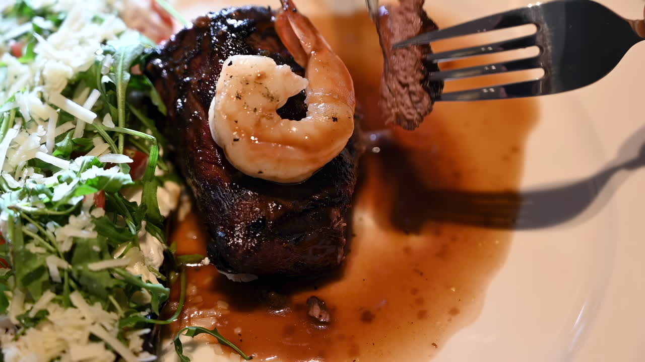 Close up of a woman cutting up a piece of beef with shrimp and a side salad