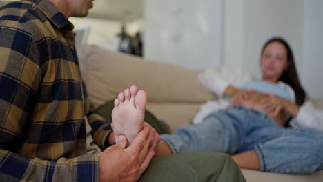 Close-up-shot-of-a-middle-aged-man-in-a-plaid-shirt-doing-a-foot-massage-for-his-wife-on-the-sofa-during-pregnancy-at-home