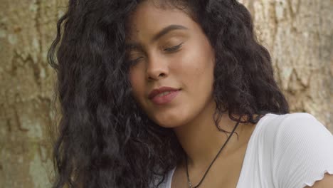 A-young-curly-hair-hispanic-girl-playing-in-her-hair-while-standing-at-a-large-tree-trunk-on-a-sunny-day