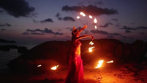 erotic and sensual belly dancer in red dress, performing with lotus shaped fire props on the beach during golden hour.